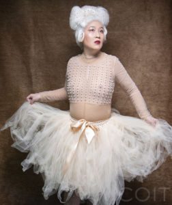 Eun Bee Yes, a nonbinary/genderqueer person in a pink tutu, pink pearl top and white wig against a brown background
