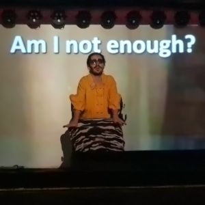 Oliver Twirl is sitting on a walker in the middle of a dark stage with the words "Am I not enough? projected behind them.